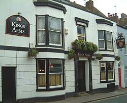 Shows a photograph of a typical old-fashioned British public house. The building is white, and it is decorated with window boxes and hanging baskets.